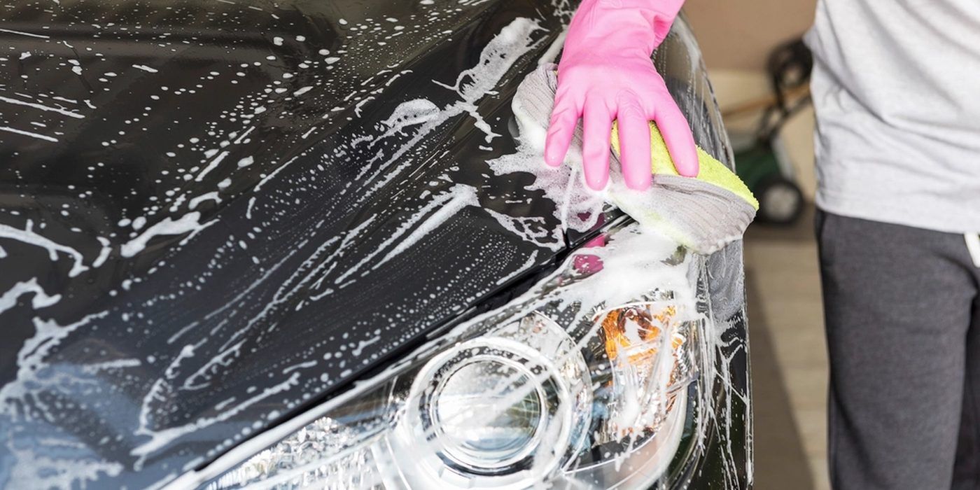 A gloved hand washing a car's headlight and bonnet under thick soap suds.
