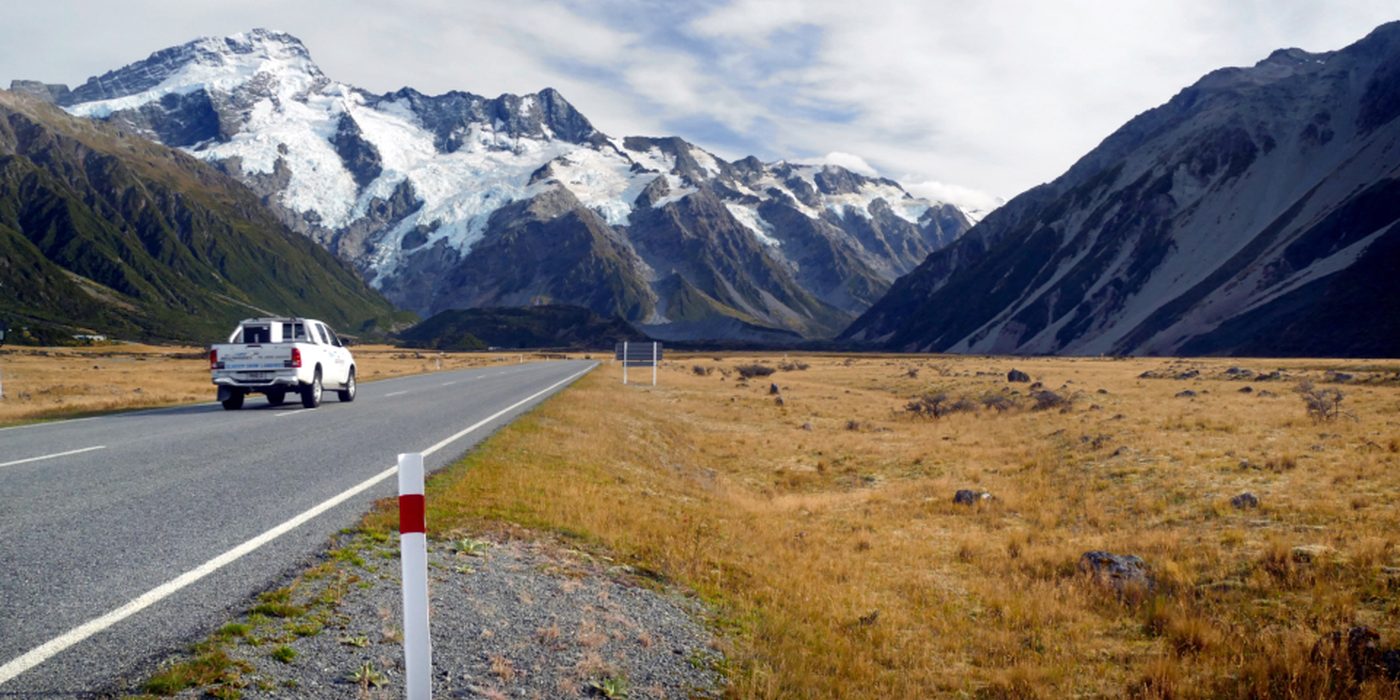 A car on an empty road running through open country towards distant mountains.