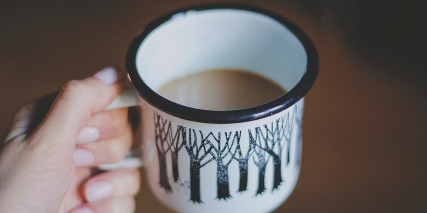 A hand holding a white enamel mug printed with a dark forest pattern.