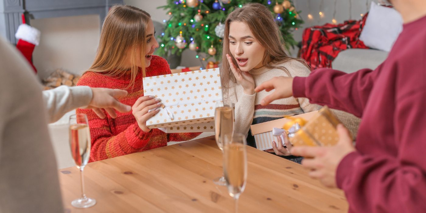 Friends in festive hats passing wrapped presents to one another across a table.