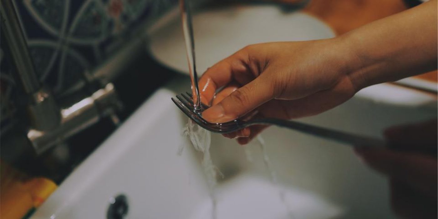 Hands rinsing cutlery under a running tap in a white kitchen sink.