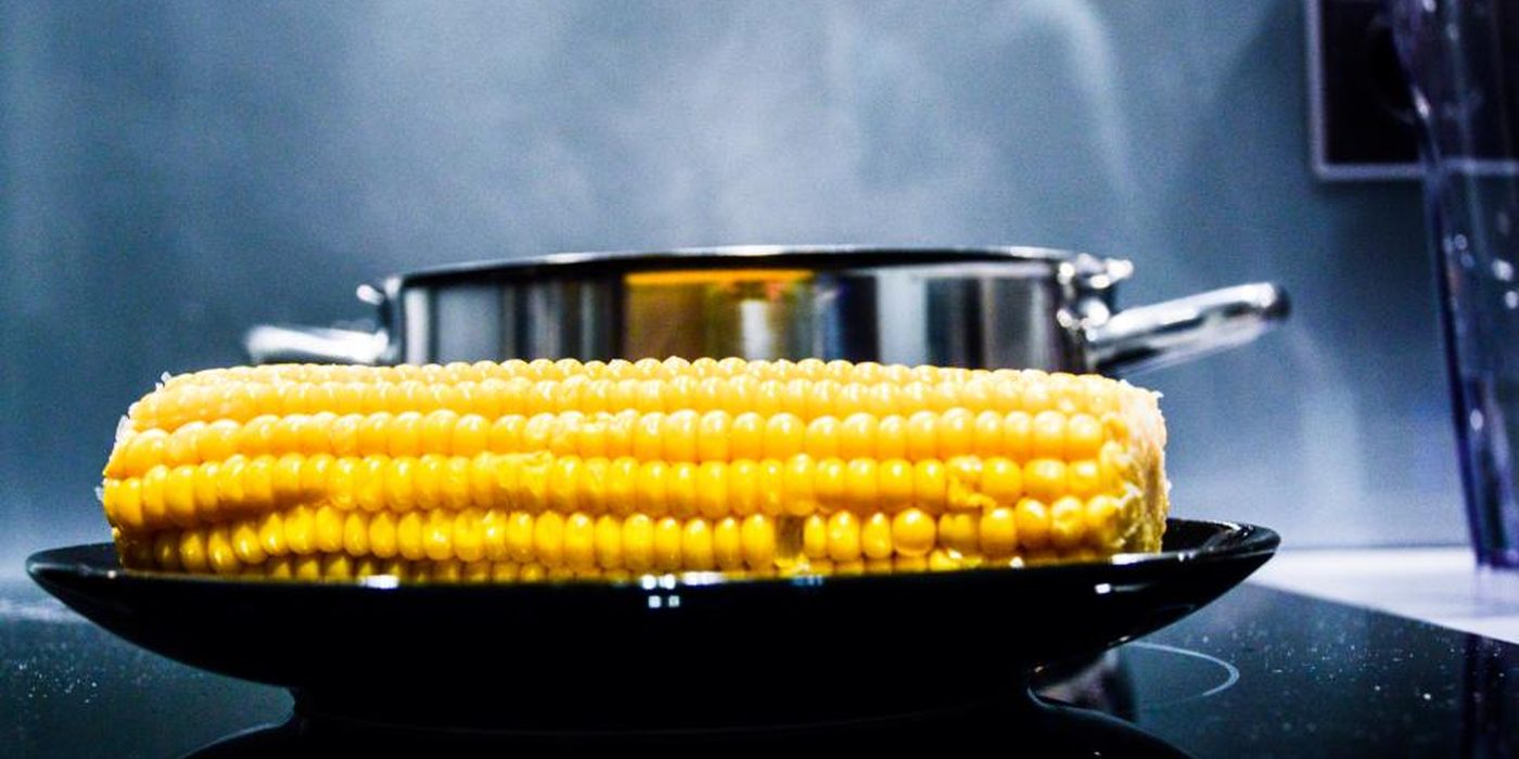 A corn cob steaming in a shallow pan on a gas stovetop, a stockpot behind it.