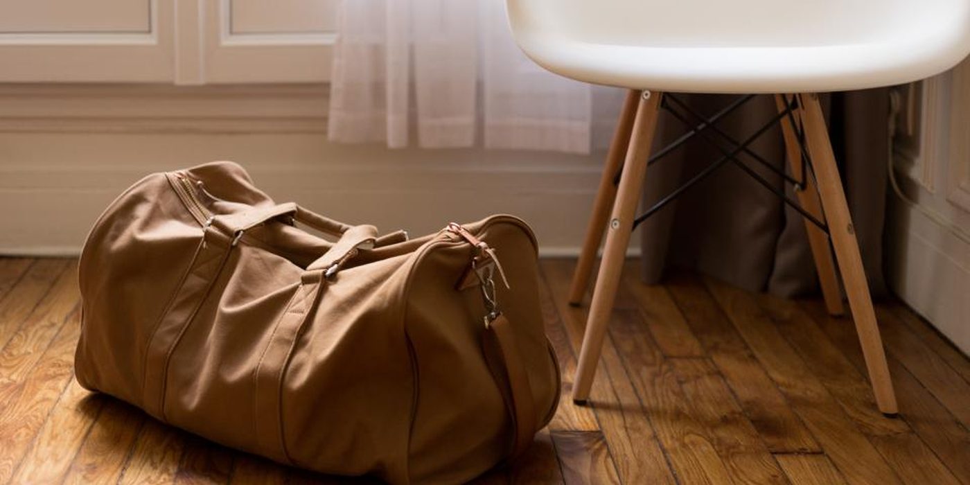 A tan leather holdall standing on a wooden floor beside a chair leg.