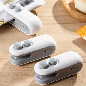 Three white and grey mini bag sealers on a wooden worktop, one of them clamped onto the top of an open snack bag