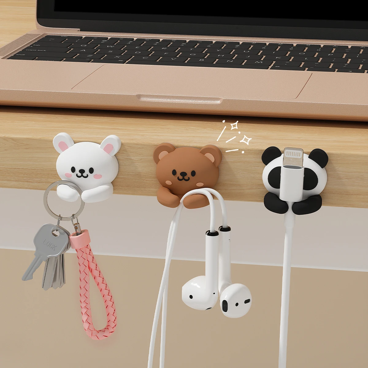 Rabbit, bear and panda cable clips on a desk edge under a laptop, holding keys, wired earphones and a charging cable