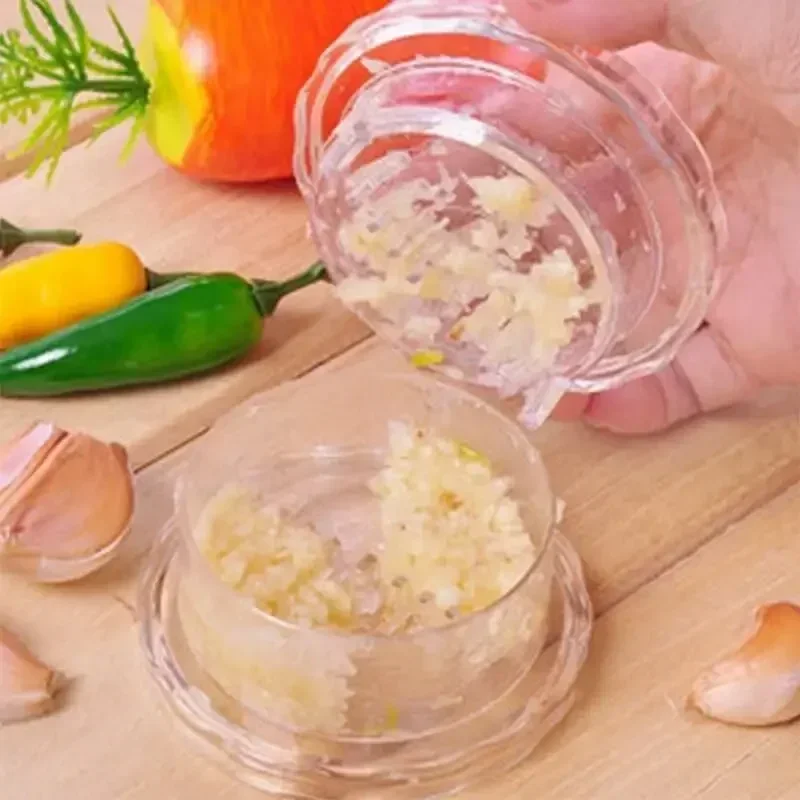 Two clear garlic crusher pots of minced garlic on a wooden board, one held tilted, with garlic cloves and peppers nearby