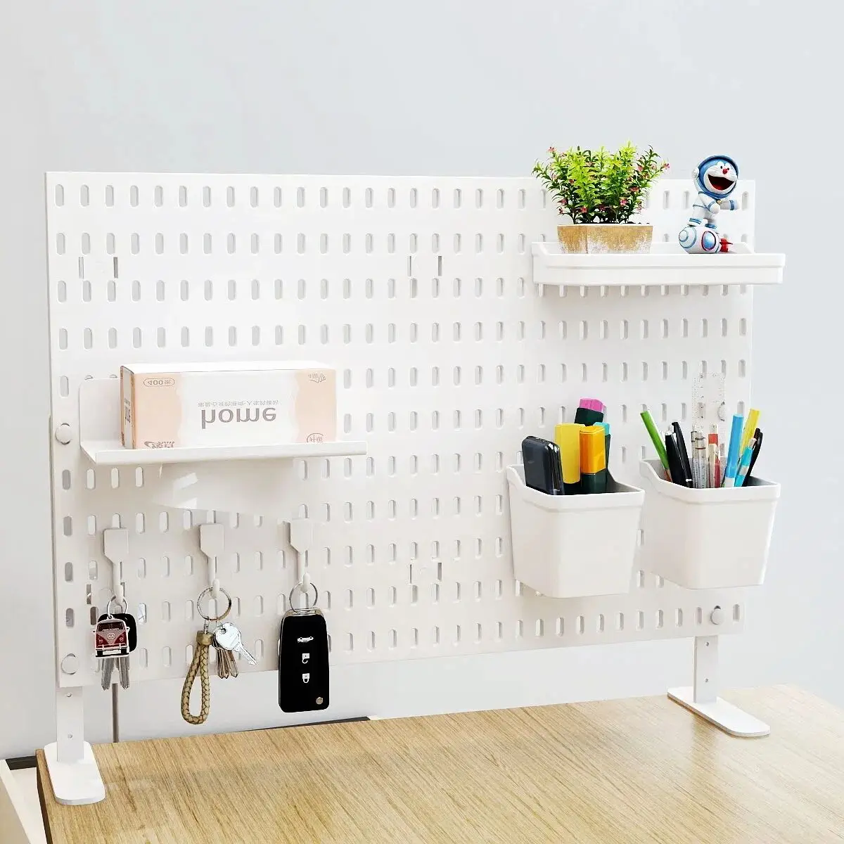 White slotted pegboard panel standing on a desk on two feet, holding two shelves, key hooks, a potted plant and two pen cups
