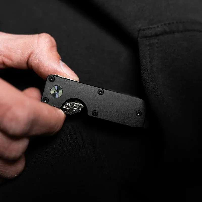 Slim black metal key organizer held closed in a hand against dark clothing, showing its pivot screw and thumb cut-out