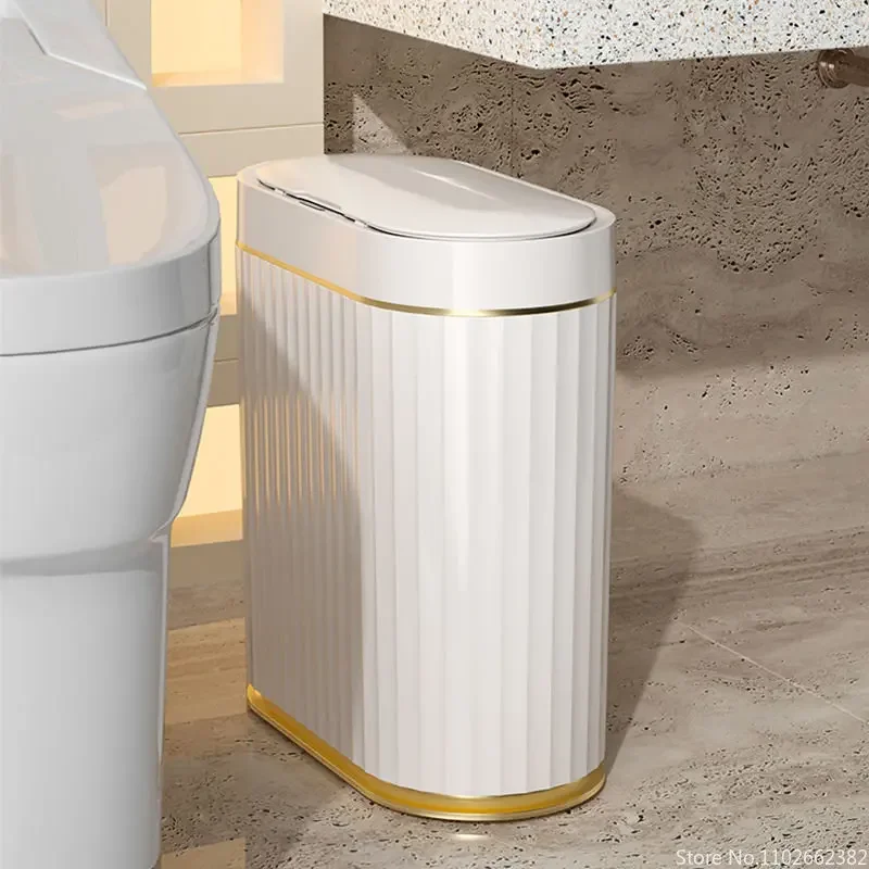 Slim white sensor bin with gold rim and base band beside a toilet in a stone-tiled bathroom, lid closed