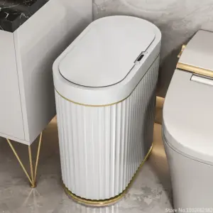 White fluted motion sensor bathroom trash can with a gold trim band, on a marble floor between a vanity and a toilet