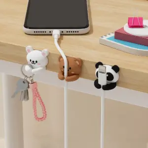 White rabbit, brown bear and panda cable clips stuck to a wooden desk edge, holding keys and a white phone charging cable