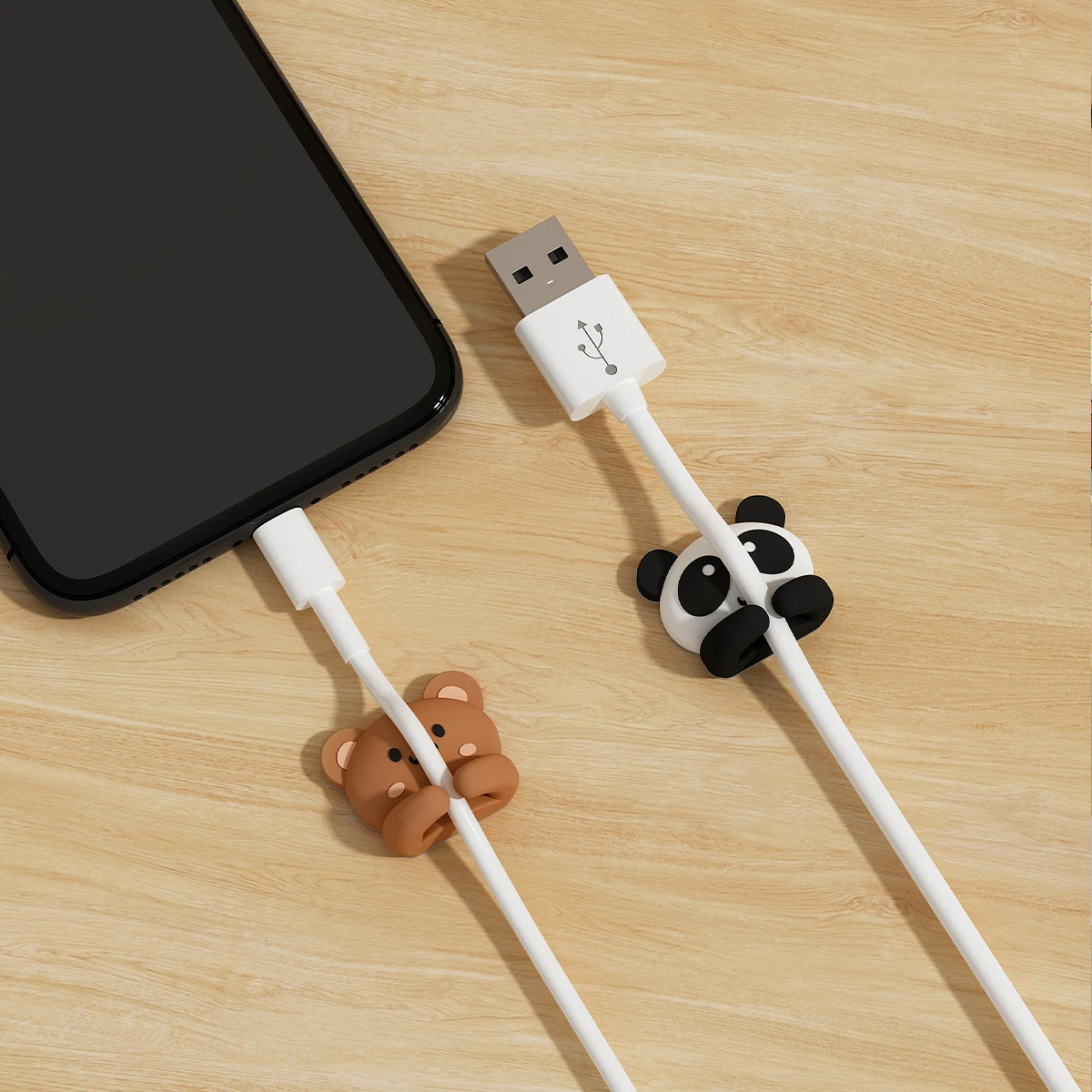 Brown bear and panda cable clips fastened part-way along a white USB charging cable beside a phone on a wooden desk